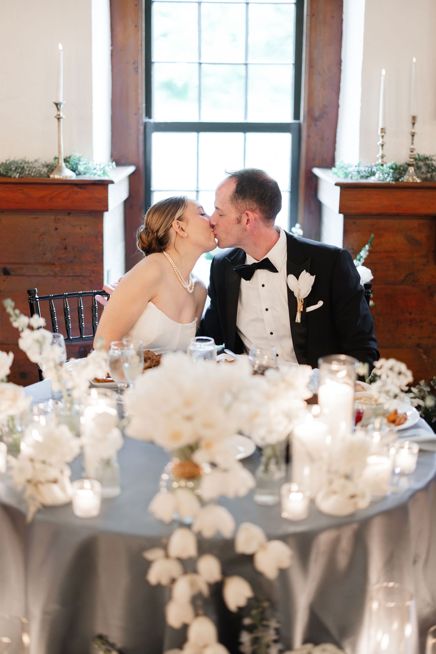 Bride and groom kiss at sweetheart table adorned with white floral centerpieces.