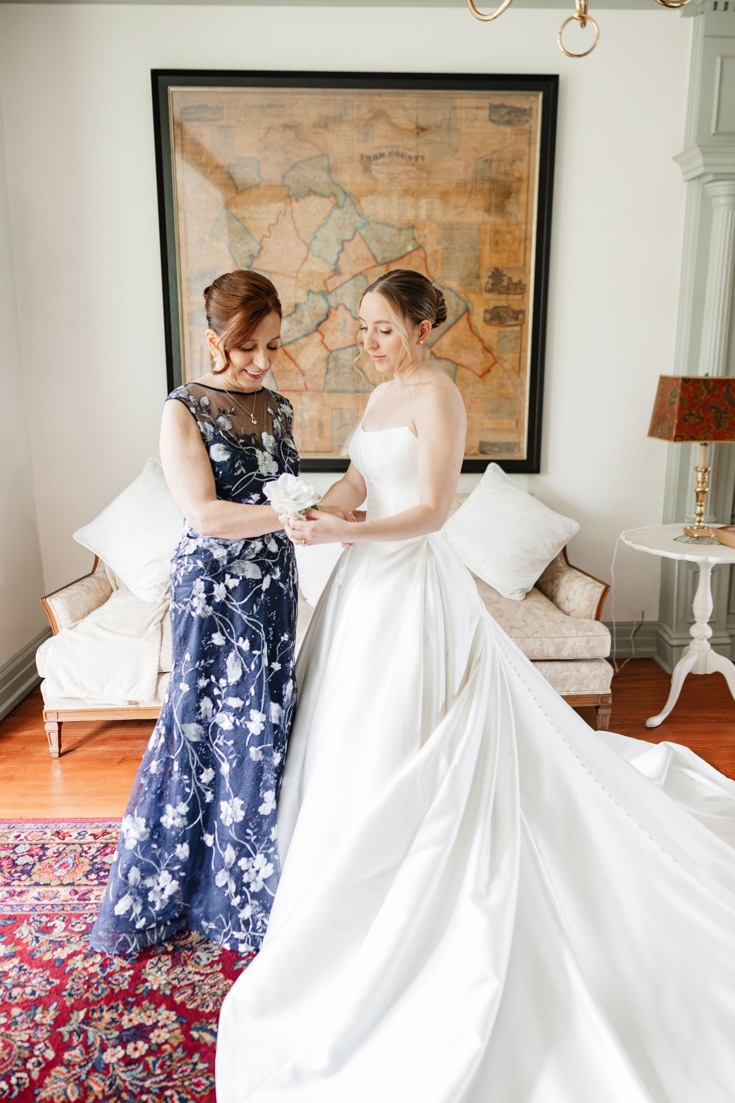 Woman in navy floral dress assists bride in white strapless ball gown in classic interior with antique map.
