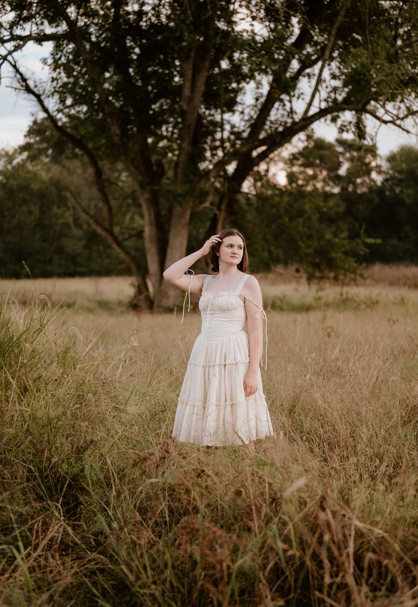 Young woman in white tiered dress standing in golden field with trees, golden hour portrait photography.