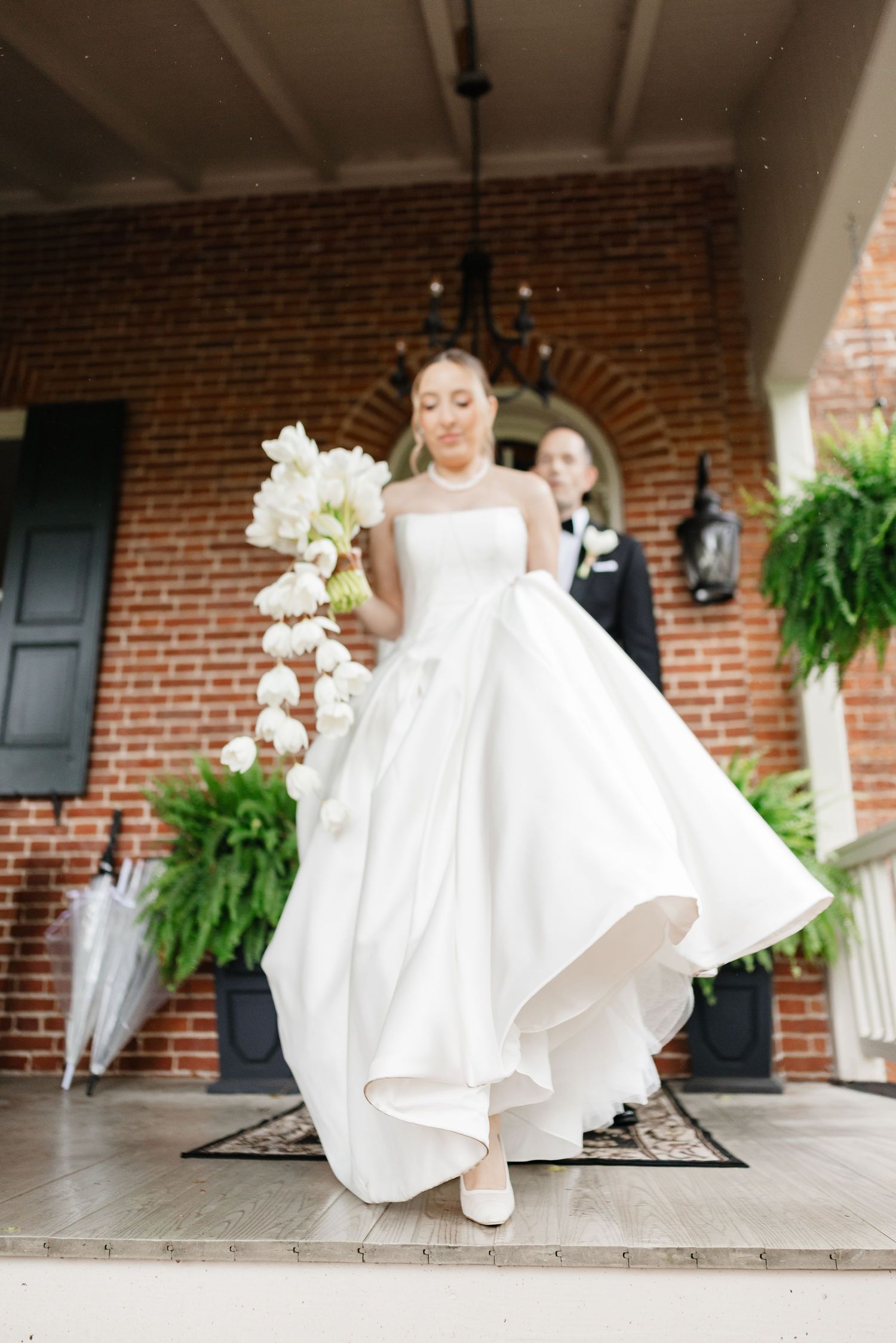 Bride in a strapless white ball gown holds a white floral bouquet while descending steps of a brick mansion.