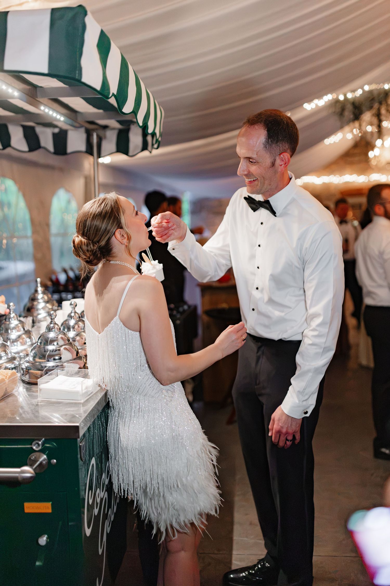 Bride in white feather dress dancing with groom in tuxedo shirt at wedding reception near elegant bar cart.