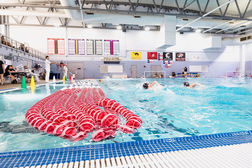 Swimmers training in indoor competition pool with red lane dividers and spectators watching.