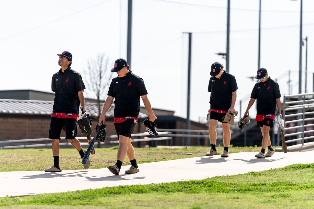Four baseball players in black uniforms walking along a path carrying equipment bags.