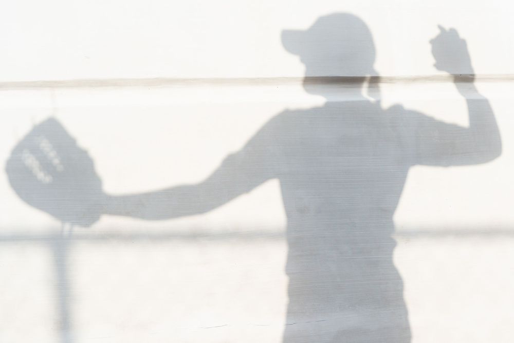 Silhouette of softball player throwing ball seen through fence in bright backlit conditions.