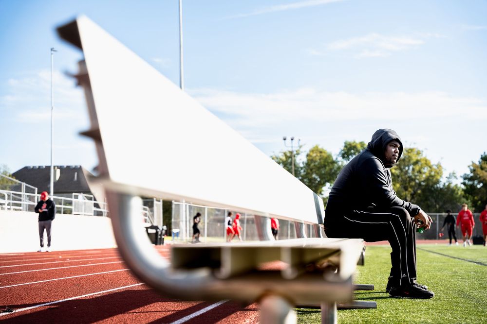 Athlete in hoodie sitting alone on sideline bench at outdoor sports track facility.