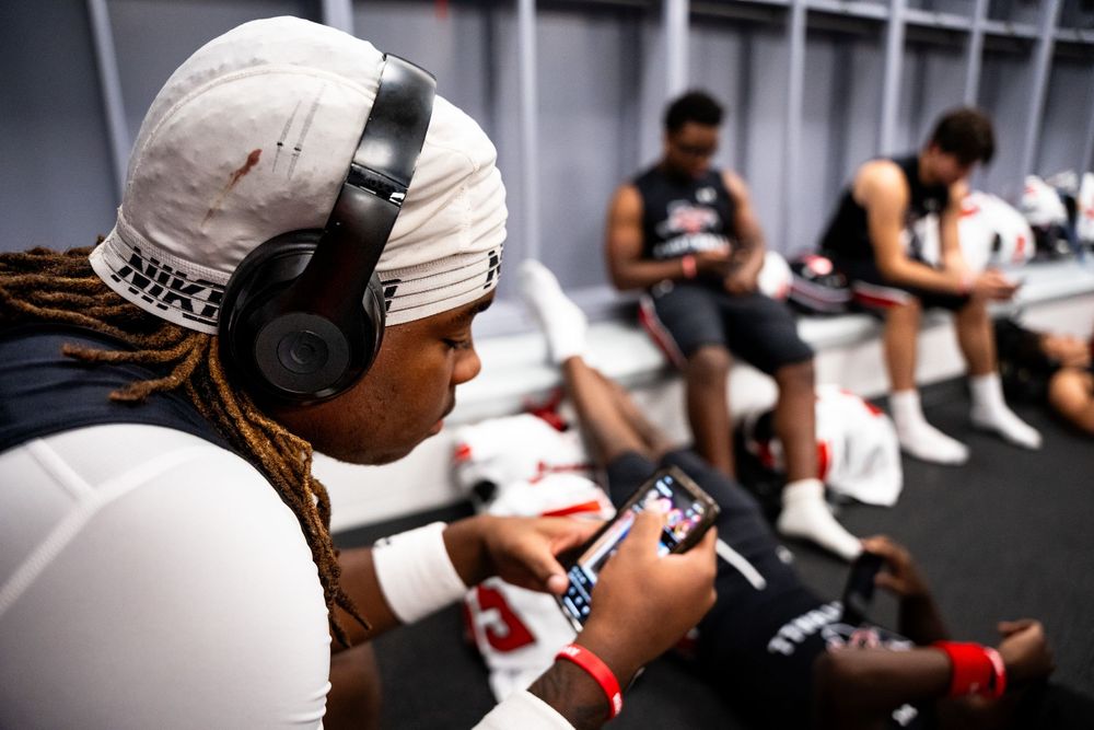 Football player wearing headphones watches video on phone in team locker room before game.