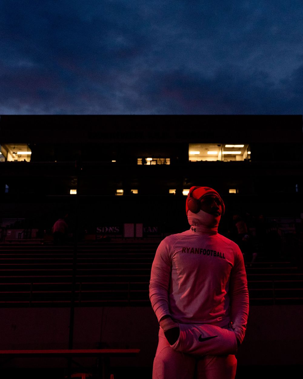 Athlete in warmup jacket stands alone at dusk before illuminated stadium grandstands.
