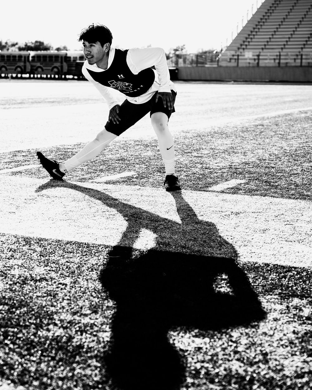 Black and white photo of football player stretching on field with dramatic shadow in stadium.