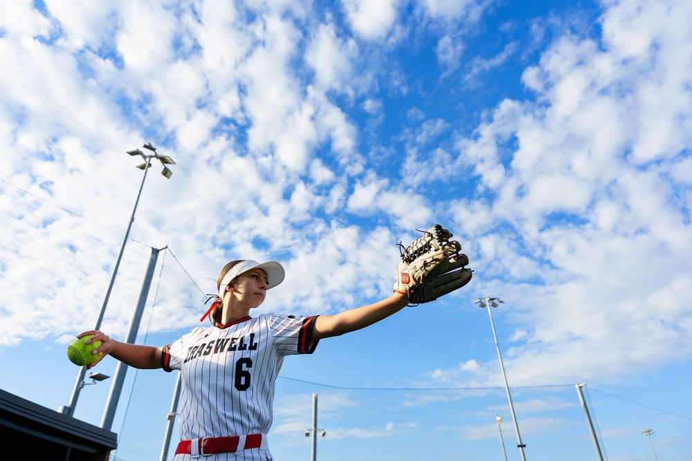 Softball player number 6 reaches up to catch ball against bright blue cloudy sky.