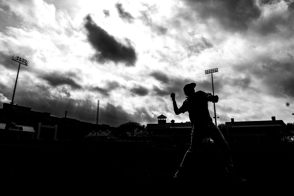 Dramatic black and white silhouette of a baseball player warming up against a cloudy sky at a stadium.