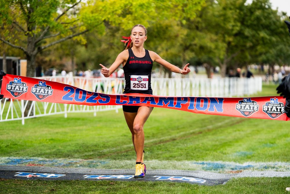 Female cross country runner breaks the finish line tape as 2025 state champion at an outdoor meet.