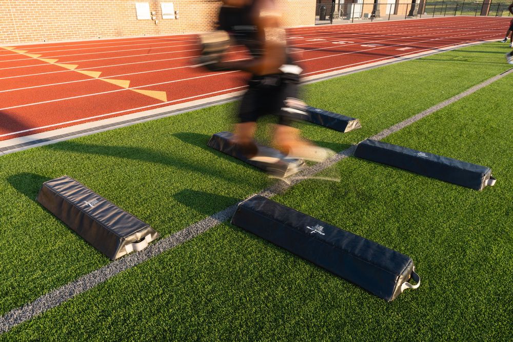 Athlete sprinting over agility training pads on an artificial turf field with a red running track.