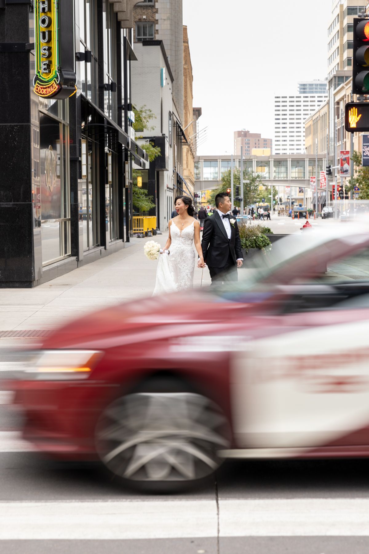 Bride and groom walk In Minneapolis city downtown in wedding attire as a blurred red car passes in the foreground.