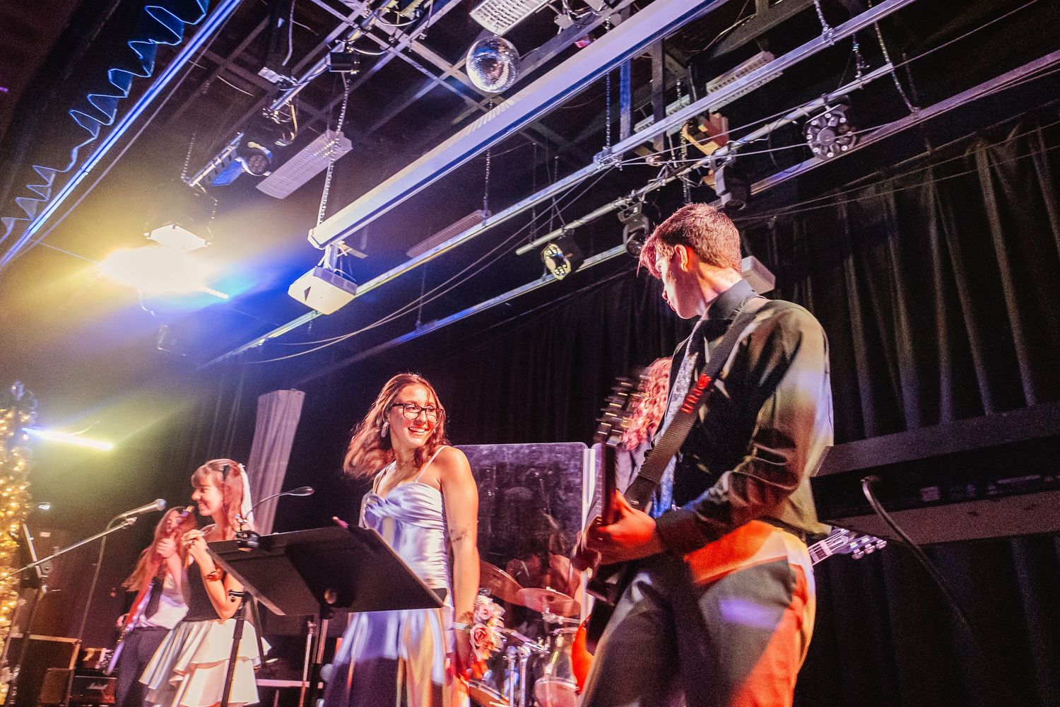 Two female singers perform on stage with a guitarist and drummer during a live band concert.
