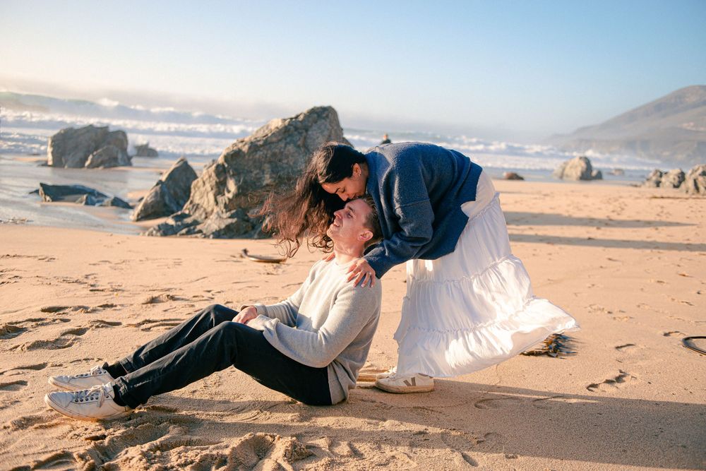 Couple sharing a romantic kiss on a sandy beach with ocean waves and rocks in the background.