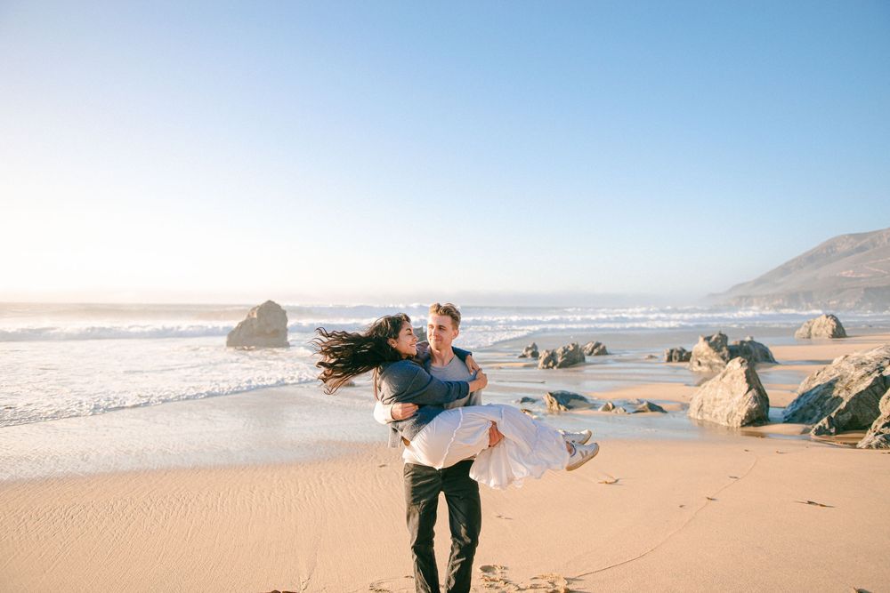 Couple embracing on a sunny beach, man lifting woman as waves crash on rocky shore behind them.