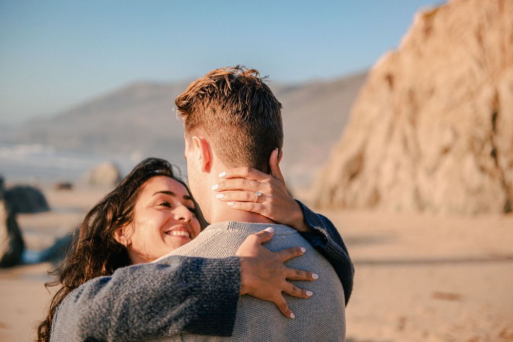 Couple embracing on a sandy beach with rocky cliffs, woman smiling joyfully in a warm, sunny coastal setting.