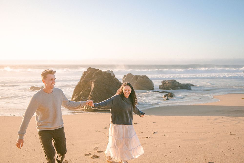A couple holds hands and walks along a sunny beach with ocean waves and rocky cliffs in the background.