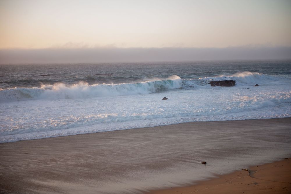 Waves crashing on a sandy beach at dusk with a misty horizon over the ocean.