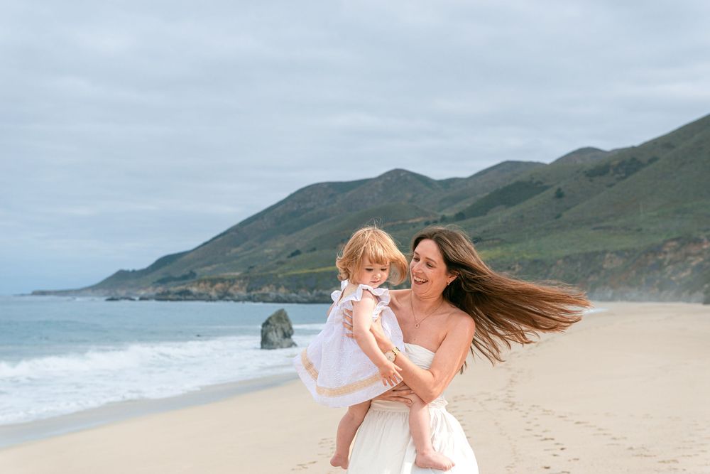 A mother holds her toddler daughter on a sandy beach with green mountains and ocean waves in the background.