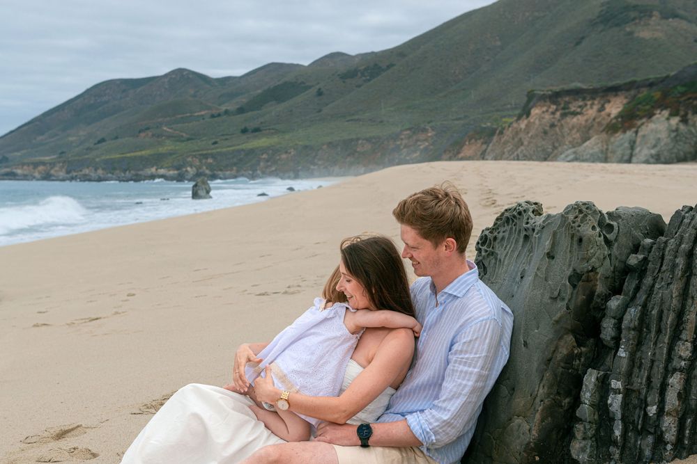 Couple sitting together on a sandy beach leaning against driftwood with scenic coastal cliffs and ocean waves in background.