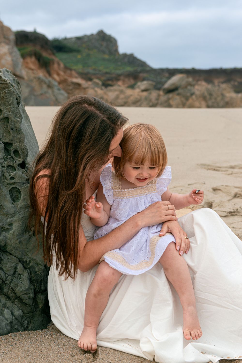 Mother in white dress holds smiling toddler girl on sandy beach with rocky cliffs in background.