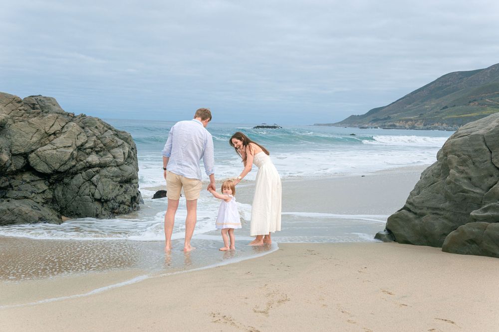 Family of three standing on a sandy beach near large rocks, with ocean waves in the background on a cloudy day.