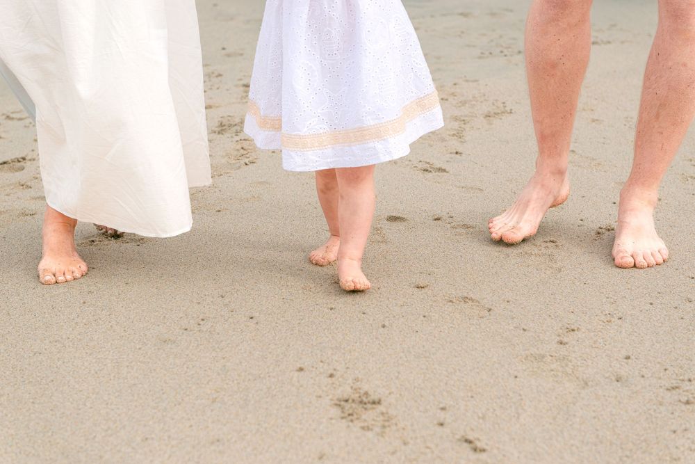 Three barefoot people walking on sandy beach, two adults in white clothing with a child in white dress.