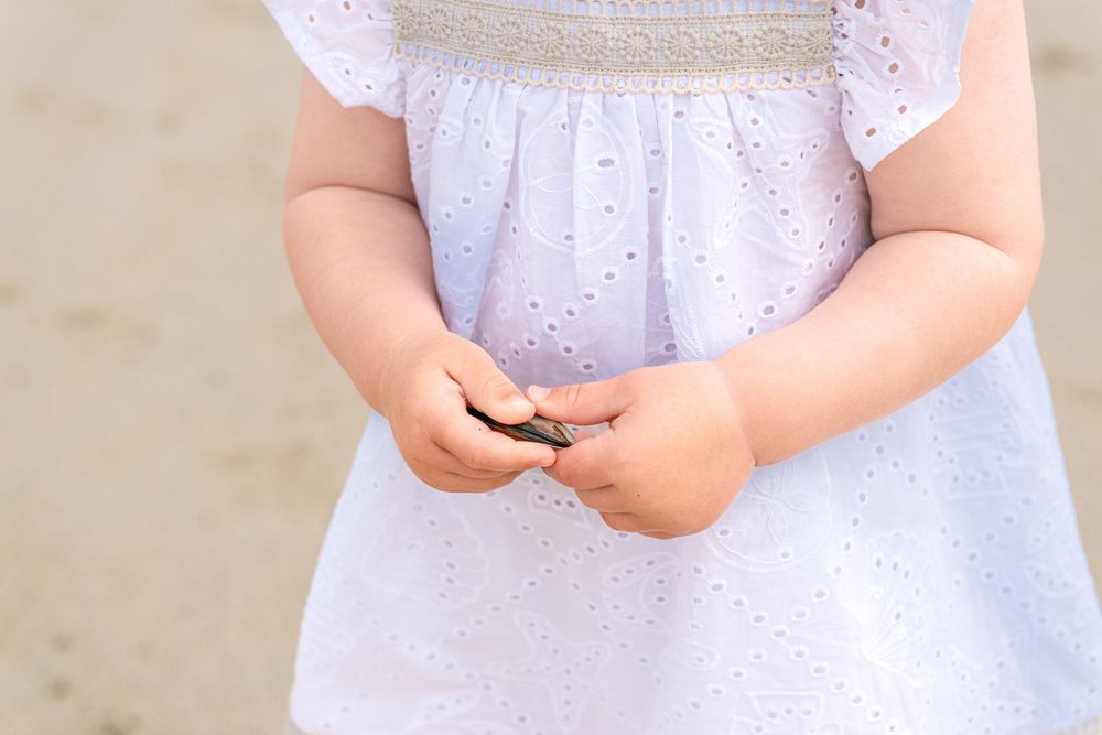 Young toddler girl in white eyelet dress holding a small rock or shell in her chubby hands outdoors.