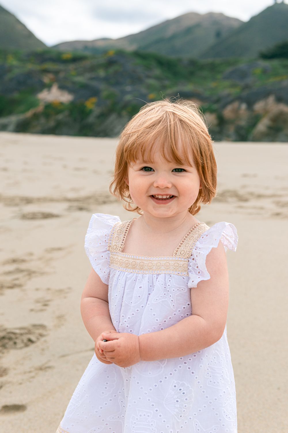 A smiling toddler girl in a white dress plays on a sandy beach with green hills in the background.