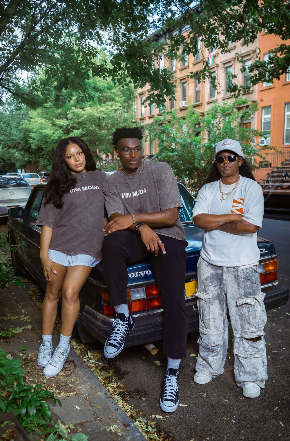 Three young people pose in front of a vintage Volvo car on a tree-lined urban street wearing casual streetwear.