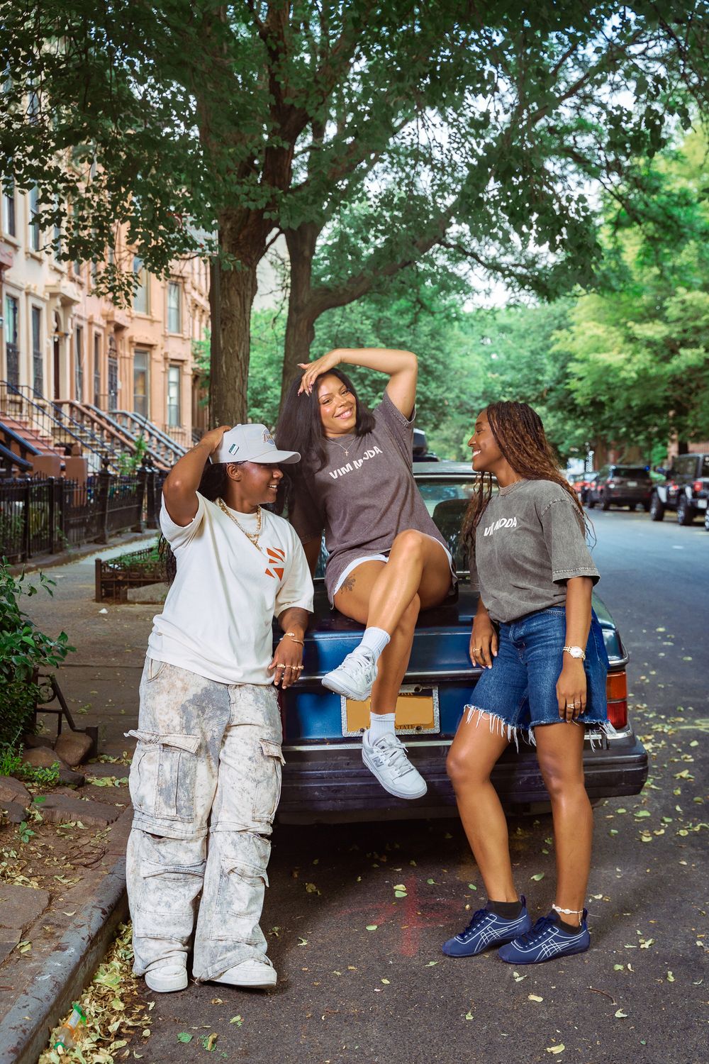 Three stylish young women posing on a city street beside a vintage blue car under a large tree.