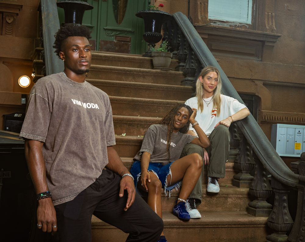Three young adults in casual streetwear pose on brownstone steps in an urban setting.