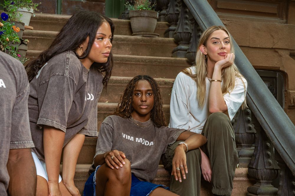 Three young women sitting on stone steps, wearing casual streetwear including graphic tees and olive pants.