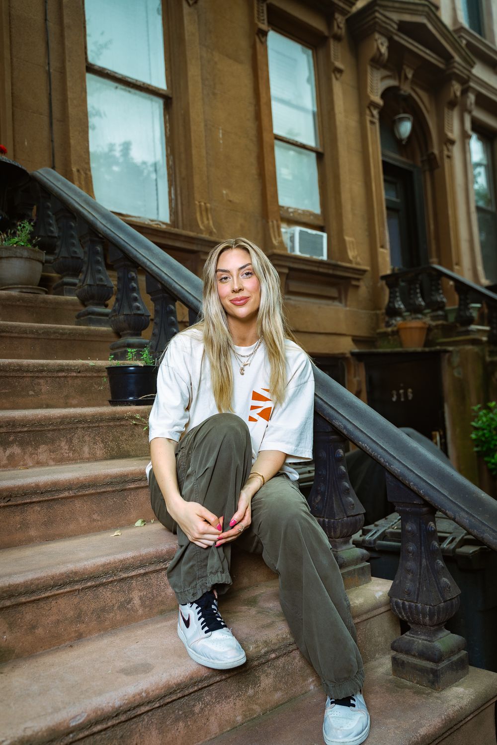 Young woman in white tee and cargo pants sitting on brownstone steps in urban NYC neighborhood.