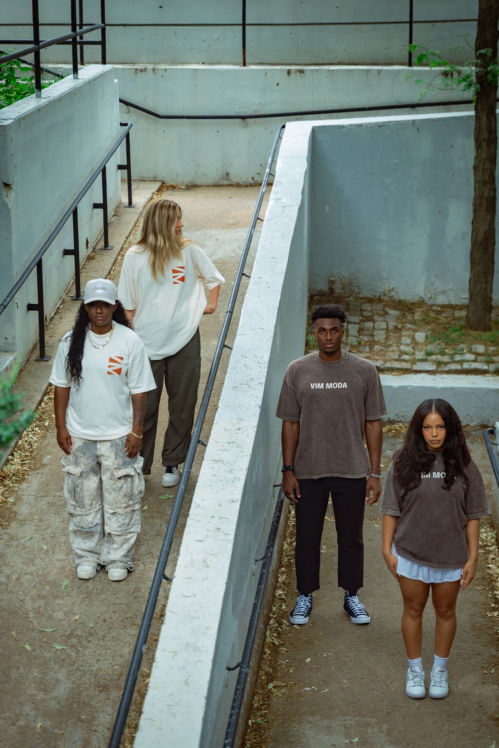 Four young people in casual streetwear pose on urban concrete steps, wearing branded oversized t-shirts and relaxed pants.