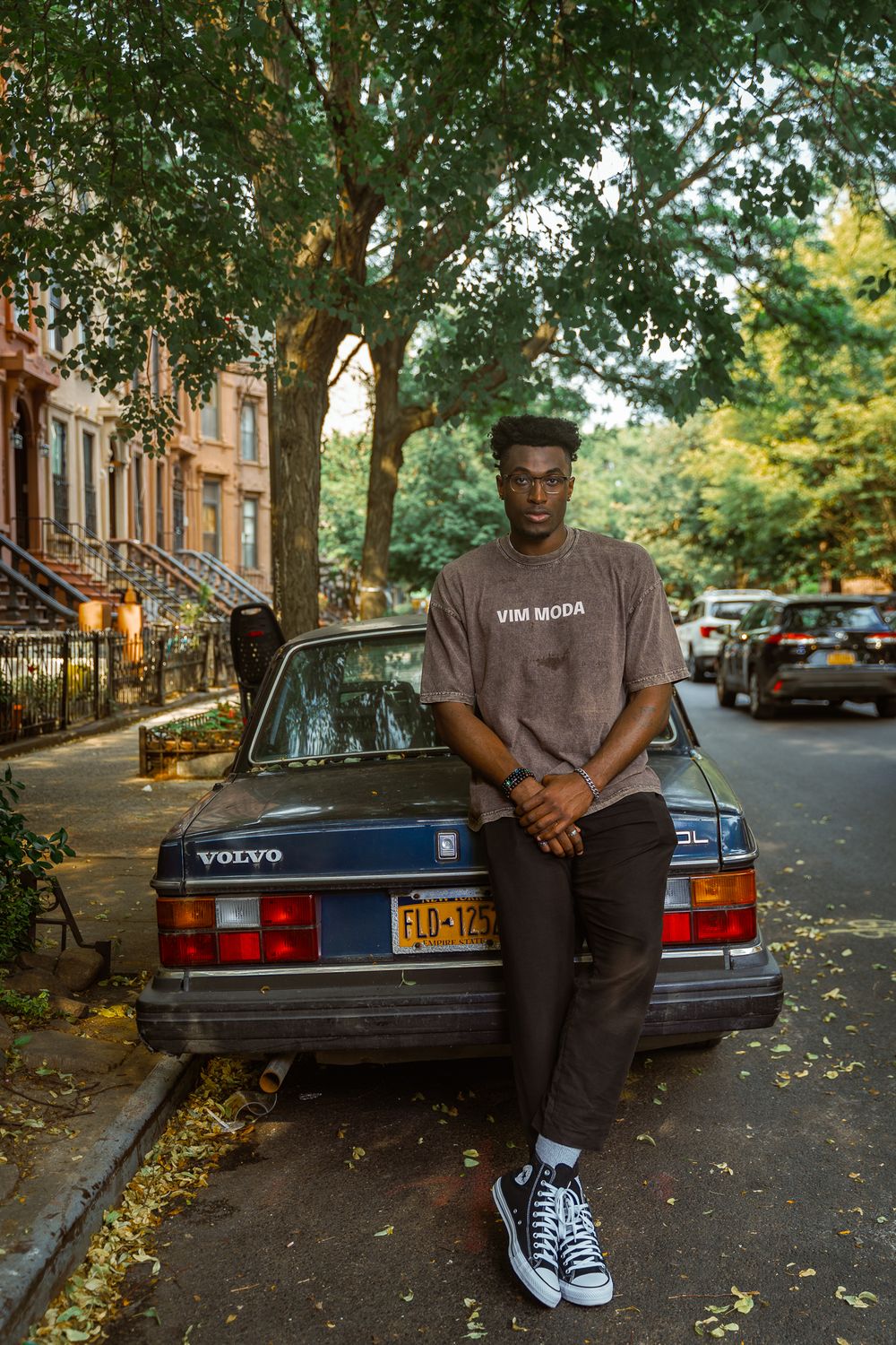 Young man in brown VIM MODA tee and Converse leans on vintage Volvo on a tree-lined Brooklyn street.