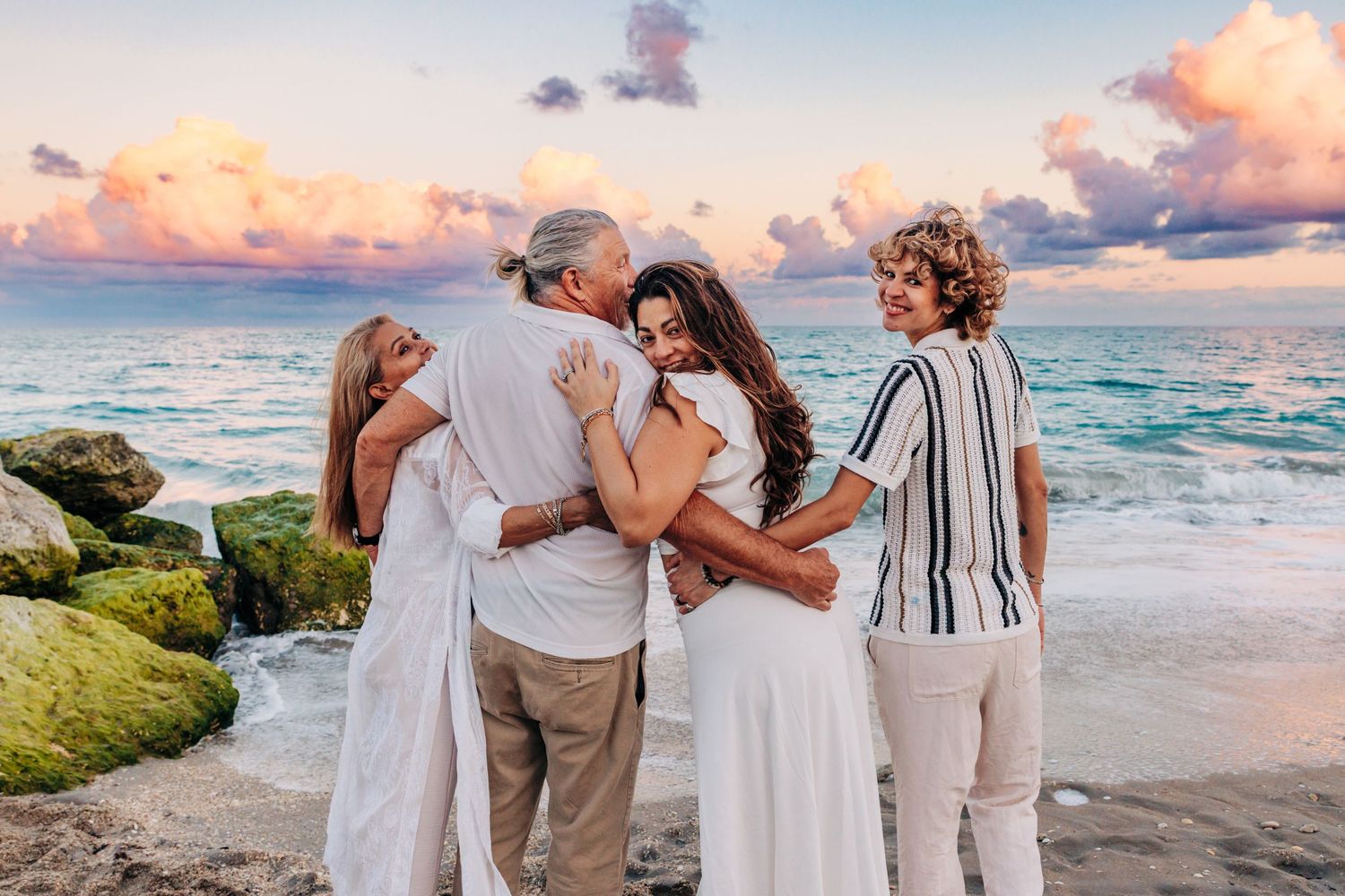 Family embracing on a rocky palm beach at sunset with colorful pink and blue sky over the ocean.