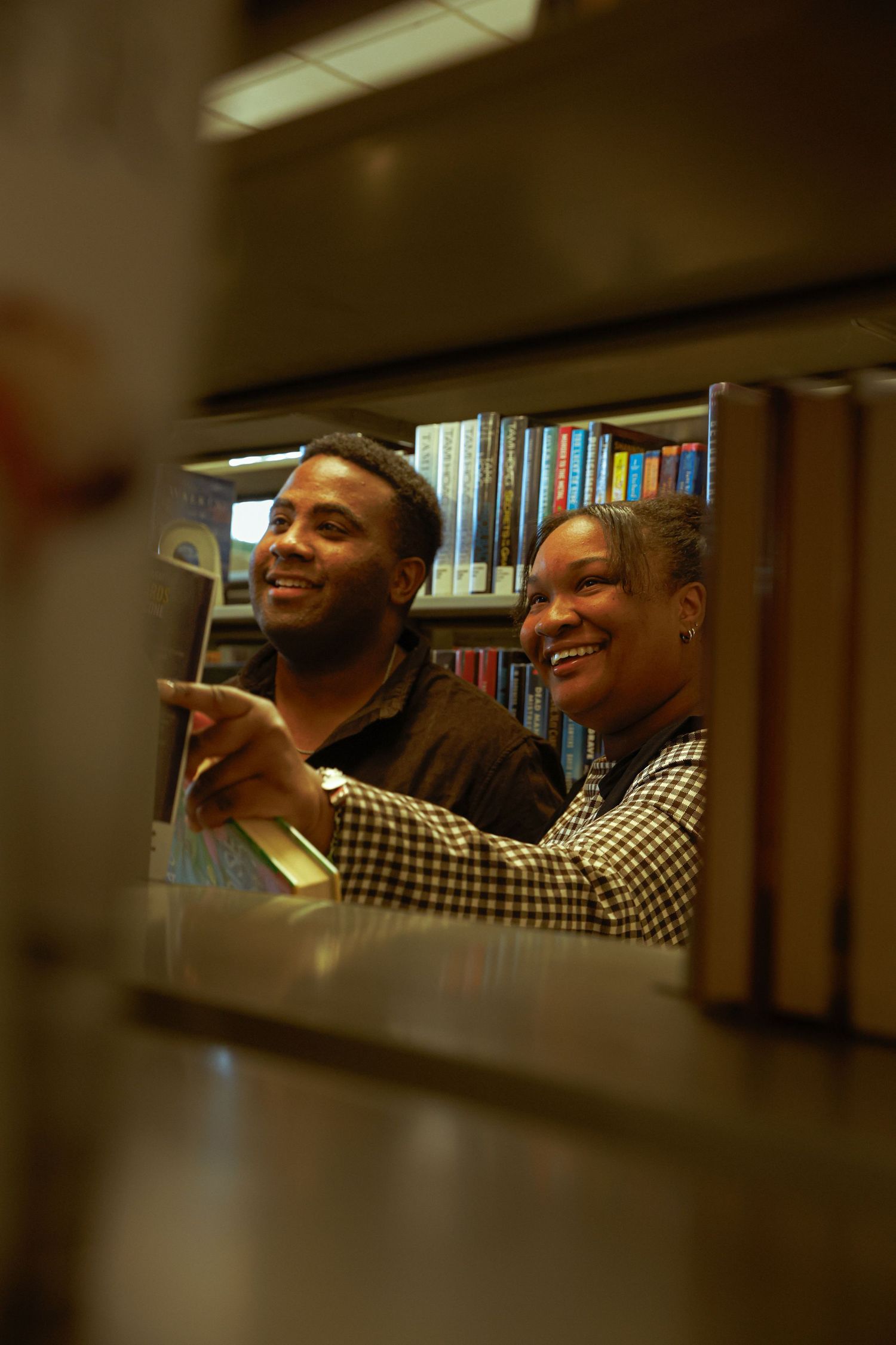 Two people smiling and browsing books together in a library, viewed through a gap in the bookshelves.