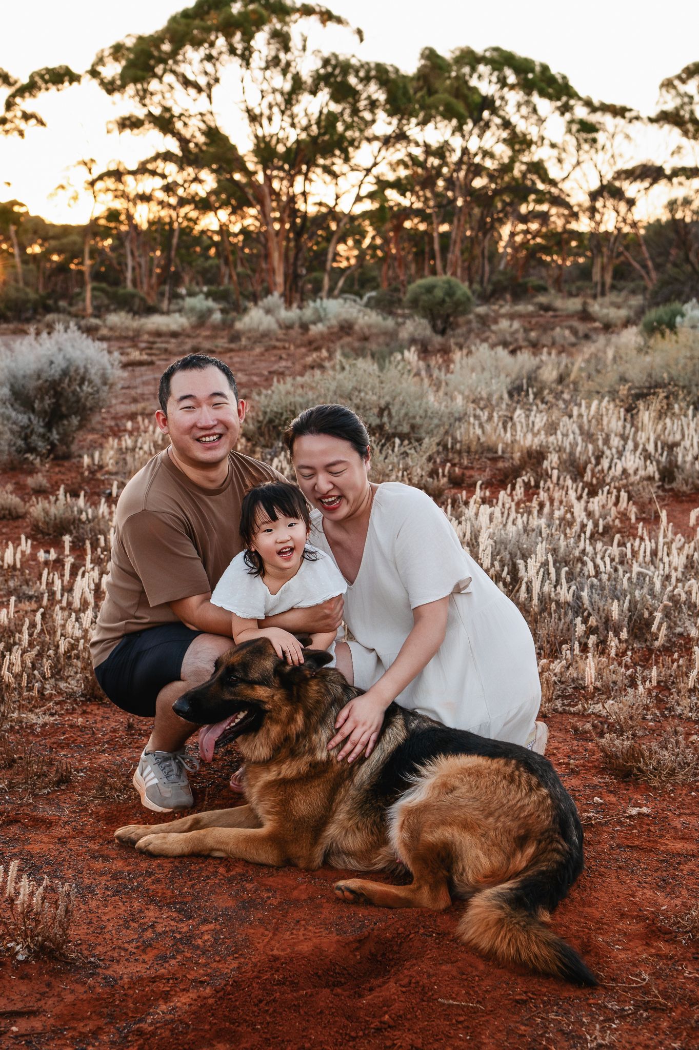 Happy Asian family of three posing outdoors in golden hour light among desert shrubs, parents holding toddler daughter.