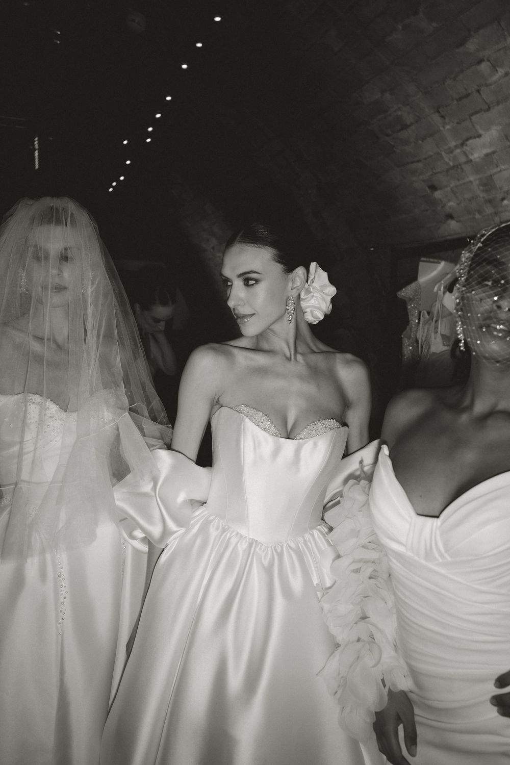 Black and white photo of models wearing elegant strapless bridal gowns, center bride adorned with floral hair accessory.
