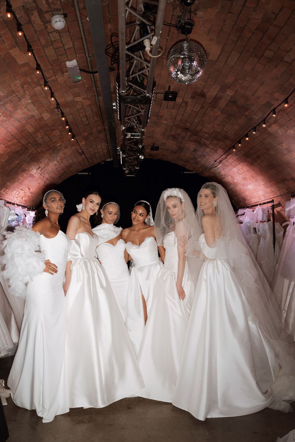 Six women pose in elegant white bridal gowns inside a brick-arched venue with warm lighting and wedding dresses displayed nearby.