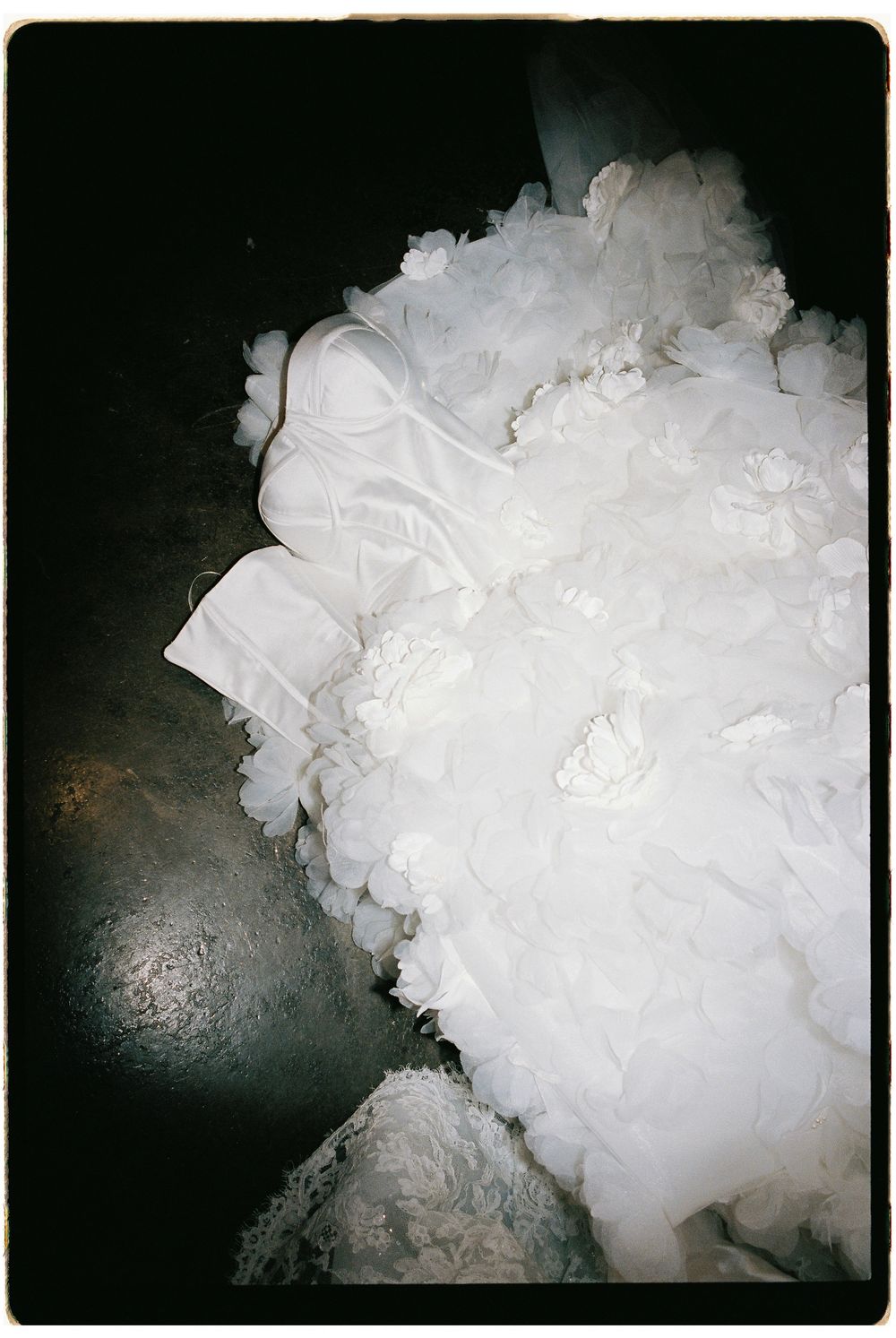 White ruffled wedding dress with floral details laying on a dark floor, photographed with vintage film aesthetic.