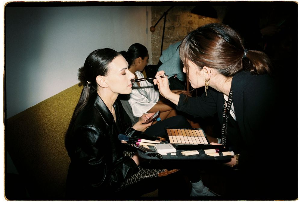 Makeup artist applying cosmetics to a model backstage, with a palette of products displayed nearby.