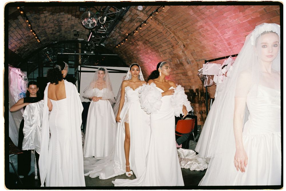 Models wearing elegant white bridal gowns and veils backstage at a fashion show in a brick-vaulted venue.