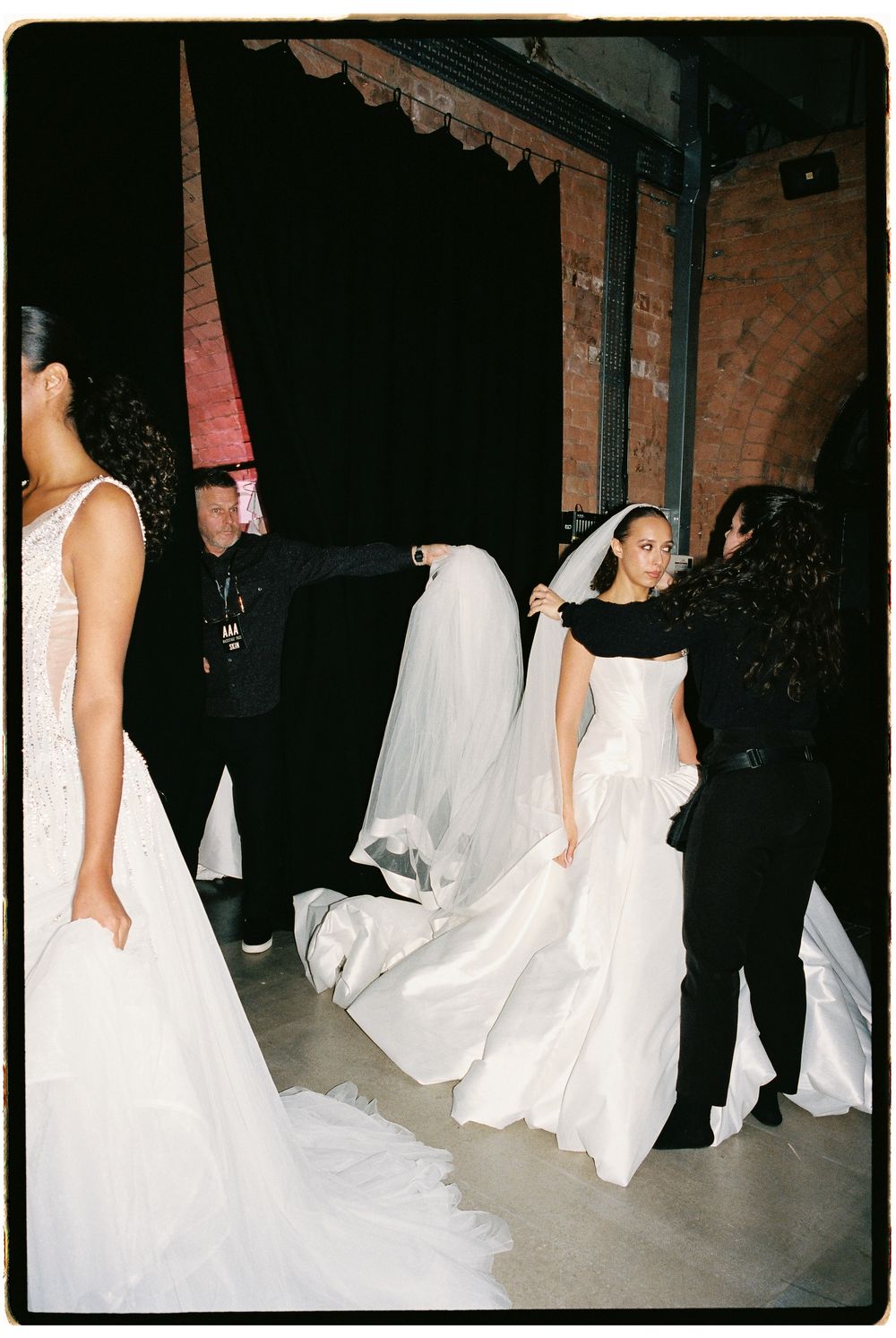 Backstage wedding fashion show with models in white bridal gowns and veils being assisted by dressers.