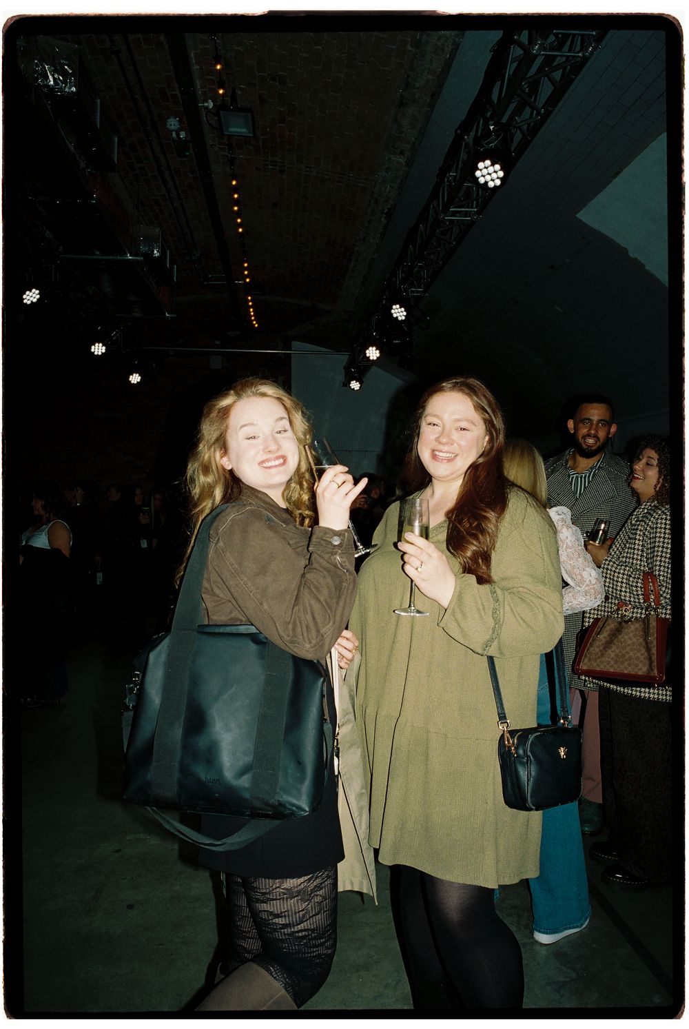 Two smiling women with handbags pose at an indoor event venue with stage lighting in the background.