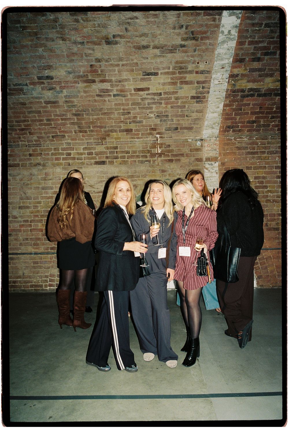 Group of women socializing at an indoor event venue with exposed brick walls, holding champagne glasses.