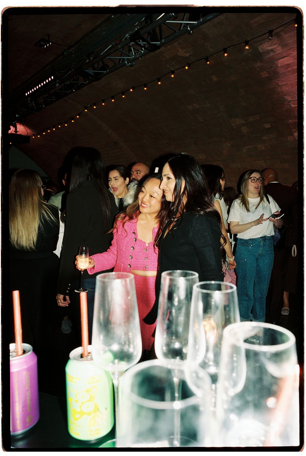 Two women embrace at a dimly lit indoor party with string lights, drinks on a table in the foreground.