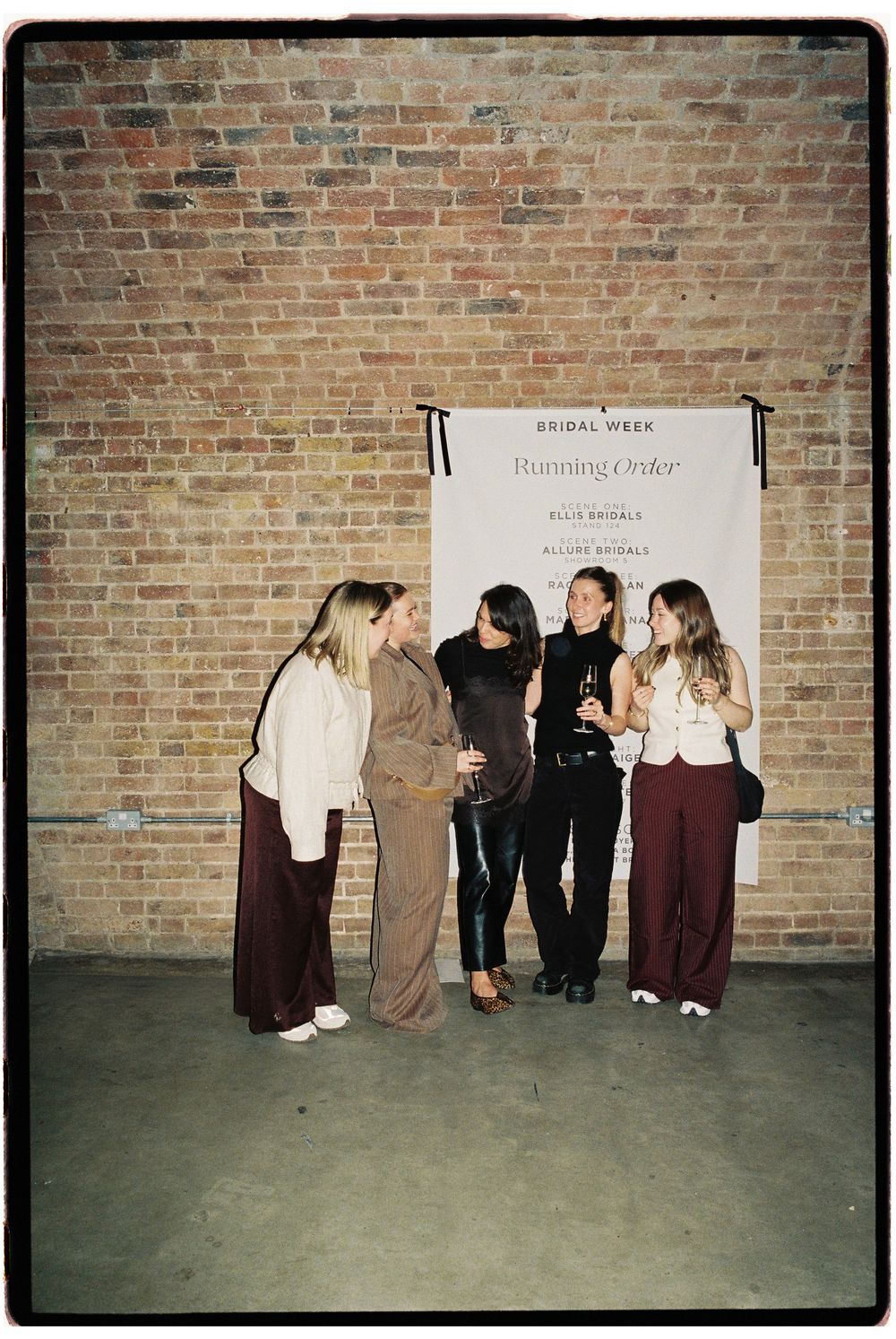 Four women socializing at a Bridal Week Running Order event in front of a brick wall backdrop.